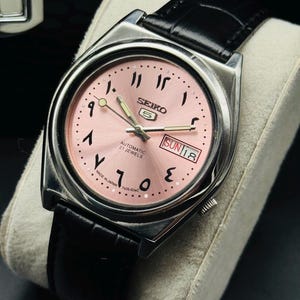 Op de afbeelding: Een zilverkleurig Seiko 5 automatisch horloge met een roze wijzerplaat en een zwarte leren band. De wijzerplaat heeft Arabische cijfers en de dag van de week. Het horloge is gemarkeerd met "Made in Japan".