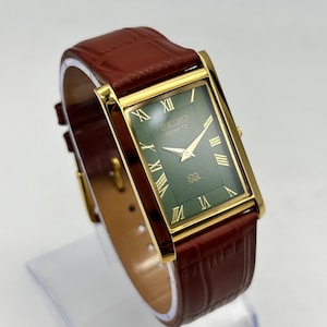 Puede incluir: Reloj Seiko Quartz dorado rectangular con esfera verde, manecillas doradas y n&uacute;meros romanos. Correa de cuero marr&oacute;n con dise&ntilde;o de cocodrilo.