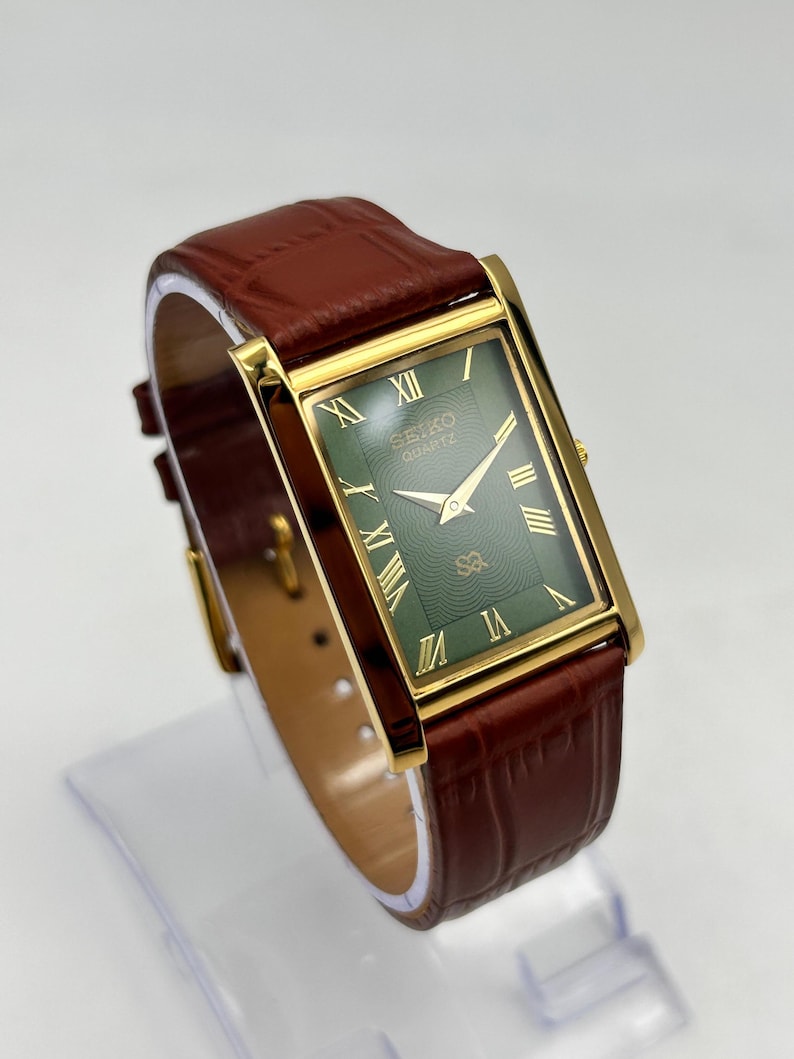 Puede incluir: Reloj Seiko Quartz dorado rectangular con esfera verde, manecillas doradas y n&uacute;meros romanos. Correa de cuero marr&oacute;n con dise&ntilde;o de cocodrilo.