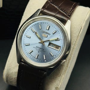 May include: A silver-toned Seiko 5 Sports watch with a light blue dial and a brown leather strap. The watch face displays the day and date, with white hour markers and hands. The watch is displayed on a cream-colored cushion.