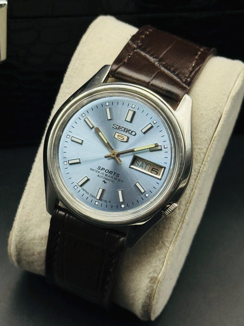 May include: A silver-toned Seiko 5 Sports watch with a light blue dial and a brown leather strap. The watch face displays the day and date, with white hour markers and hands. The watch is displayed on a cream-colored cushion.