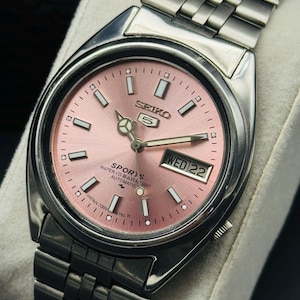 May include: A stainless steel Seiko 5 Sports automatic watch with a pink dial. The watch features a day and date display, luminous hands and markers, and a stainless steel bracelet. The dial reads "SEIKO 5 SPORTS WATER 10 BAR RESIST AUTOMATIC".