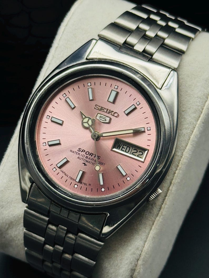 May include: A stainless steel Seiko 5 Sports automatic watch with a pink dial. The watch features a day and date display, luminous hands and markers, and a stainless steel bracelet. The dial reads "SEIKO 5 SPORTS WATER 10 BAR RESIST AUTOMATIC".