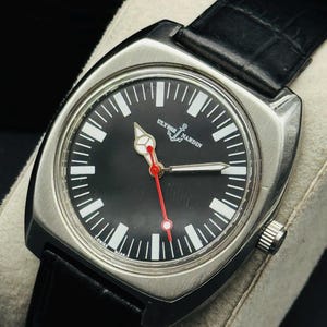 Pode incluir: Um rel&oacute;gio Ulysse Nardin em a&ccedil;o inoxid&aacute;vel com mostrador preto, marcadores de hora brancos e ponteiro de segundos vermelho. O rel&oacute;gio tem uma caixa quadrada e uma pulseira de couro preta. O mostrador exibe o nome da marca e um logotipo de &acirc;ncora.