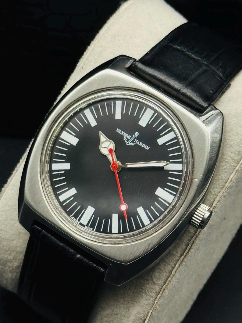Pode incluir: Um rel&oacute;gio Ulysse Nardin em a&ccedil;o inoxid&aacute;vel com mostrador preto, marcadores de hora brancos e ponteiro de segundos vermelho. O rel&oacute;gio tem uma caixa quadrada e uma pulseira de couro preta. O mostrador exibe o nome da marca e um logotipo de &acirc;ncora.