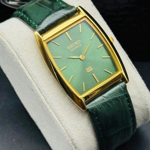 May include: A gold-toned Seiko Quartz watch with a rectangular green face and gold hands. The watch has a green leather band. The watch is displayed on a white cushion.