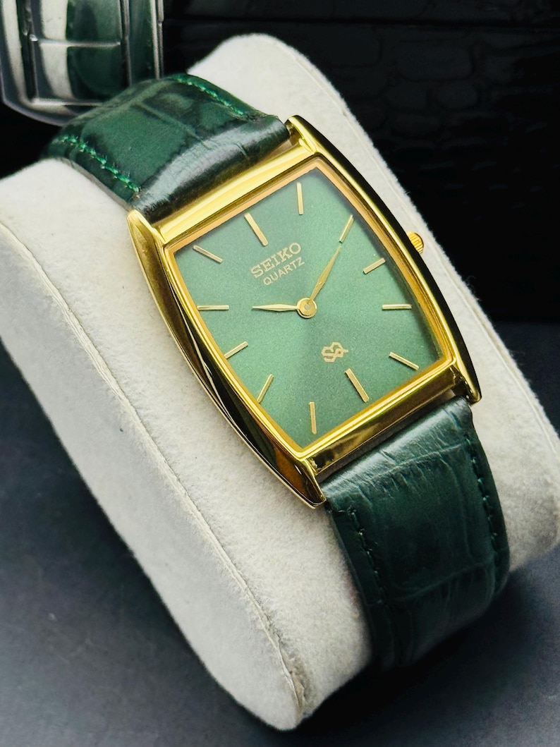 May include: A gold-toned Seiko Quartz watch with a rectangular green face and gold hands. The watch has a green leather band. The watch is displayed on a white cushion.