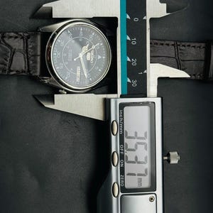 May include: A silver-toned watch with a dark gray face and a black leather band is measured with a digital caliper. The caliper displays a measurement of 35.37 mm, or approximately 1.39 inches.