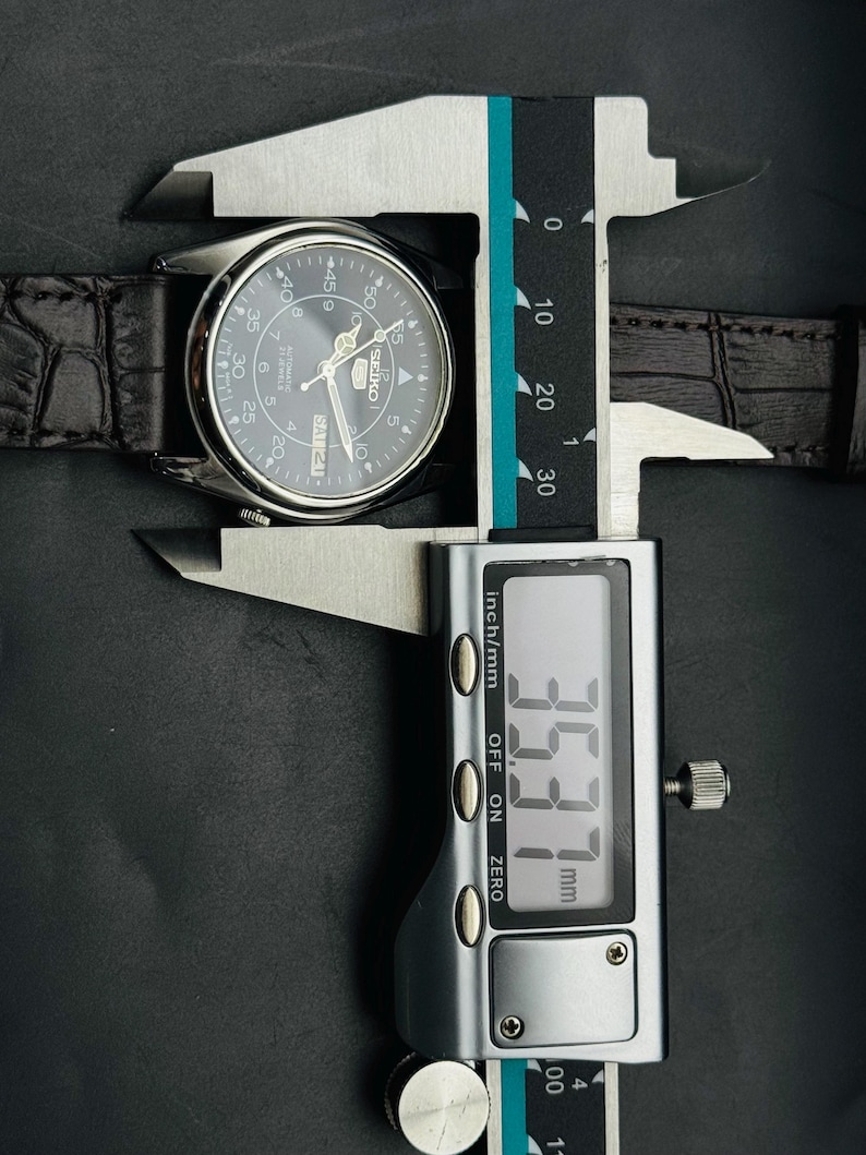 May include: A silver-toned watch with a dark gray face and a black leather band is measured with a digital caliper. The caliper displays a measurement of 35.37 mm, or approximately 1.39 inches.