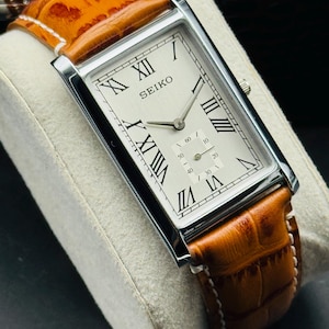 Op de afbeelding: Een rechthoekig Seiko horloge met een zilveren kast en een cr&egrave;mekleurige wijzerplaat met zwarte Romeinse cijfers. Het horloge heeft een bruine leren band met witte stiksels. Het horloge wordt gepresenteerd op een wit, getextureerd oppervlak.