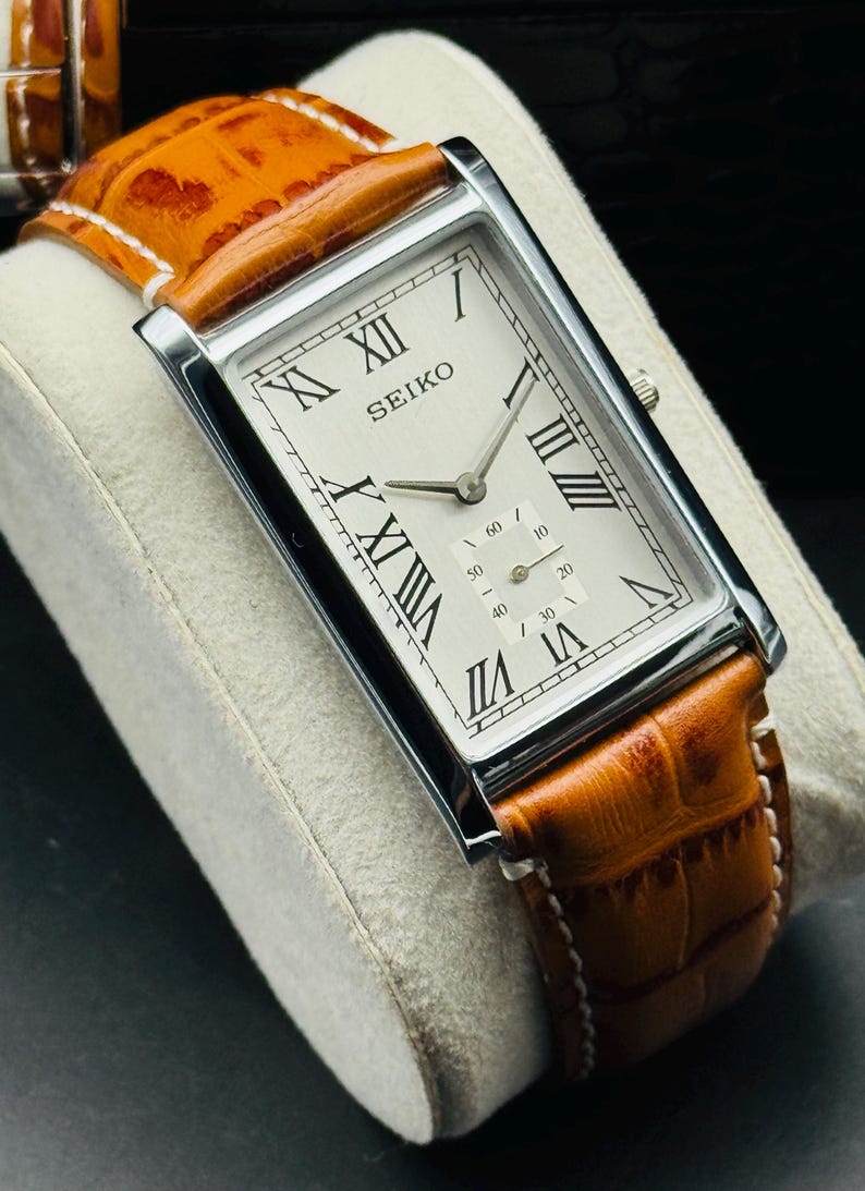 Op de afbeelding: Een rechthoekig Seiko horloge met een zilveren kast en een cr&egrave;mekleurige wijzerplaat met zwarte Romeinse cijfers. Het horloge heeft een bruine leren band met witte stiksels. Het horloge wordt gepresenteerd op een wit, getextureerd oppervlak.