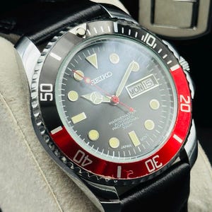 May include: A close-up of a Seiko Marine Master automatic professional watch with a black dial, a red and black bezel, and a black leather strap. The watch face displays the day and date.