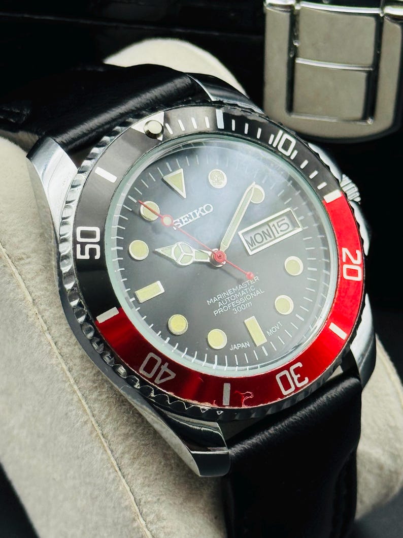 May include: A close-up of a Seiko Marine Master automatic professional watch with a black dial, a red and black bezel, and a black leather strap. The watch face displays the day and date.
