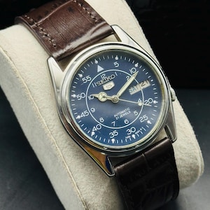 May include: A vintage Seiko 5 automatic watch with a dark blue dial and silver-tone case. The watch features a brown leather strap and displays the day and date. The dial includes Arabic numerals and luminous hands.