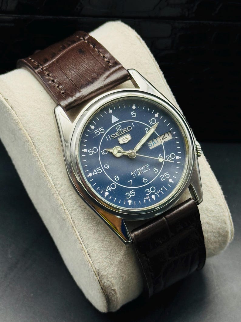 May include: A vintage Seiko 5 automatic watch with a dark blue dial and silver-tone case. The watch features a brown leather strap and displays the day and date. The dial includes Arabic numerals and luminous hands.