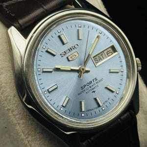 May include: A silver-toned Seiko 5 Sports watch with a light blue dial and a brown leather strap. The watch face displays the day and date, with the day reading "SAT 5". The watch has a water resistance feature.
