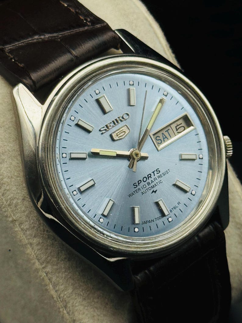 May include: A silver-toned Seiko 5 Sports watch with a light blue dial and a brown leather strap. The watch face displays the day and date, with the day reading "SAT 5". The watch has a water resistance feature.