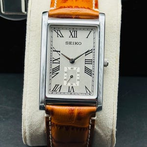 Op de afbeelding: Een rechthoekig Seiko-polshorloge met een zilverkleurige kast en een lichtgekleurde wijzerplaat met zwarte Romeinse cijfers. Het horloge heeft een bruine leren band met witte stiksels. Het horloge wordt gepresenteerd op een cr&egrave;mekleurig kussen.