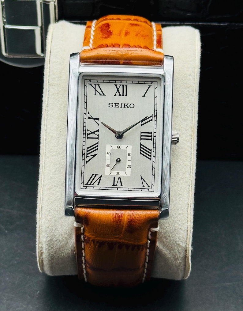 Op de afbeelding: Een rechthoekig Seiko-polshorloge met een zilverkleurige kast en een lichtgekleurde wijzerplaat met zwarte Romeinse cijfers. Het horloge heeft een bruine leren band met witte stiksels. Het horloge wordt gepresenteerd op een cr&egrave;mekleurig kussen.