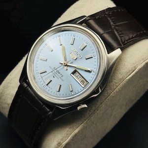 May include: A silver-toned Seiko 5 Sports watch with a light blue dial and a brown leather strap. The watch face displays the day and date. The watch is on a light gray pillow.