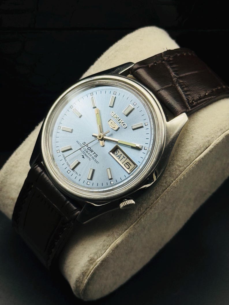 May include: A silver-toned Seiko 5 Sports watch with a light blue dial and a brown leather strap. The watch face displays the day and date. The watch is on a light gray pillow.