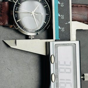 May include: A silver-toned wristwatch with a gray face and brown leather band is measured with a digital caliper. The watch face displays the brand name and the words "SWISS MADE". The caliper reads 38.16 mm.