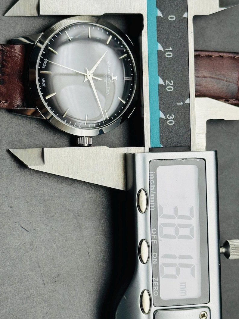 May include: A silver-toned wristwatch with a gray face and brown leather band is measured with a digital caliper. The watch face displays the brand name and the words "SWISS MADE". The caliper reads 38.16 mm.