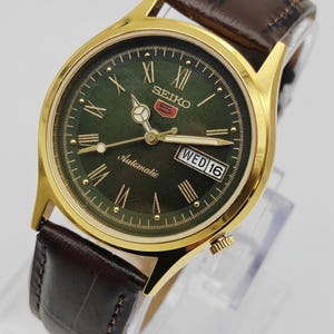 Könnte beinhalten: Eine goldfarbene Seiko 5 Automatik-Uhr mit dunkelgrünem Zifferblatt, römischen Ziffern und Datumsanzeige. Die Uhr hat ein braunes Lederarmband. Das Zifferblatt zeigt das Seiko-Logo und das Wort "Automatic".