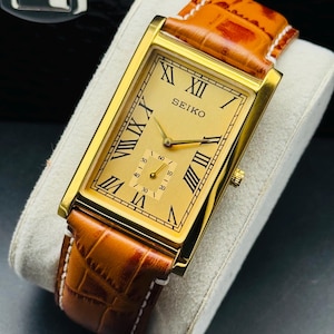 Könnte beinhalten: Eine goldfarbene Seiko-Armbanduhr mit rechteckigem Zifferblatt und römischen Ziffern. Die Uhr hat ein goldenes Zifferblatt, ein braunes Lederarmband mit weißen Nähten und eine kleine Sekundenscheibe. Die Uhr wird auf einem weißen Kissen präsentiert.