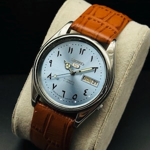 Op de afbeelding: Een zilveren Seiko 5 automatisch horloge met een lichtblauwe wijzerplaat en Arabische cijfers. Het horloge heeft een bruine leren band en toont "MON 21". Gemaakt in Japan.