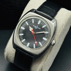 Pode incluir: Um rel&oacute;gio Ulysse Nardin em a&ccedil;o inoxid&aacute;vel com mostrador preto, marcadores brancos e ponteiro de segundos vermelho. O rel&oacute;gio tem uma pulseira de couro preta e &eacute; exibido em uma almofada cinza.