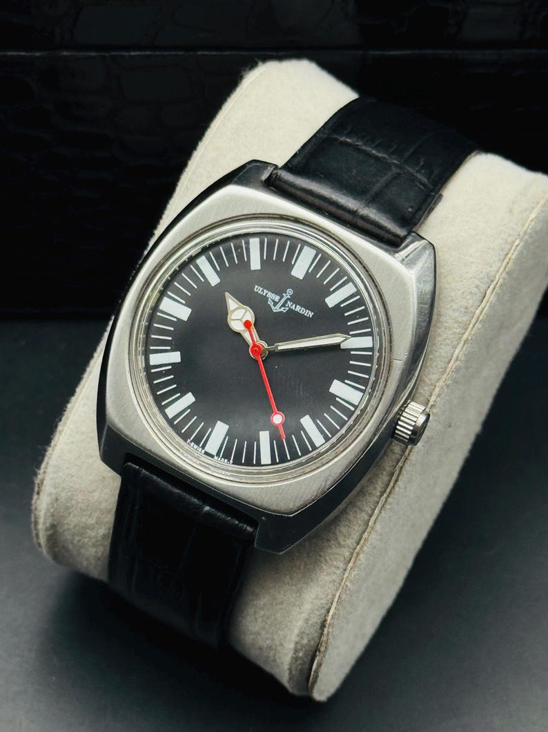 Pode incluir: Um rel&oacute;gio Ulysse Nardin em a&ccedil;o inoxid&aacute;vel com mostrador preto, marcadores brancos e ponteiro de segundos vermelho. O rel&oacute;gio tem uma pulseira de couro preta e &eacute; exibido em uma almofada cinza.