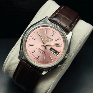 Könnte beinhalten: Eine Vintage Seiko 5 Sports Automatik-Uhr mit rosa Zifferblatt und braunem Lederarmband. Die Uhr hat ein silberfarbenes Gehäuse und zeigt Tag und Datum an. Die Uhr wird auf einem weißen Kissen präsentiert.