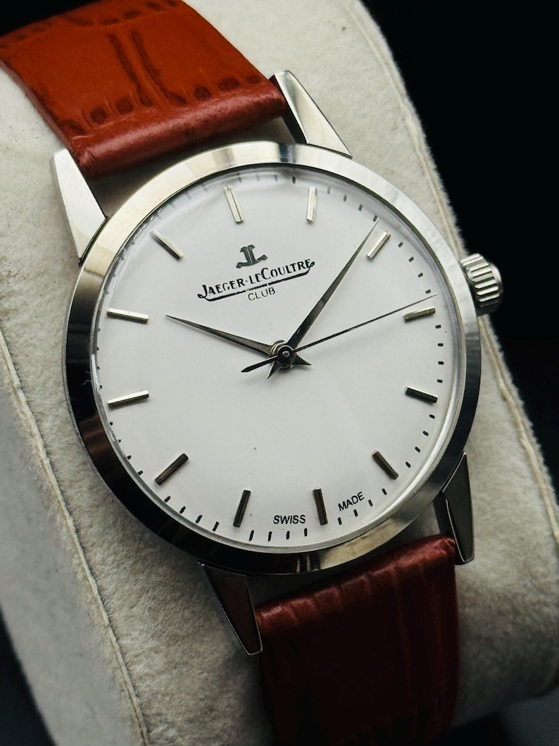 Puede incluir: Un reloj Jaeger-LeCoultre Club plateado con esfera blanca y correa de cuero marr&oacute;n. El reloj presenta agujas e indicadores plateados, y la esfera muestra el logotipo de Jaeger-LeCoultre. La correa es de un color marr&oacute;n rojizo intenso.