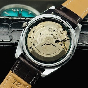 May include: Close-up of a vintage wristwatch with a silver case and a brown leather strap. The watch's back is transparent, revealing the intricate mechanical movement. The strap is marked "LEATHER 18".