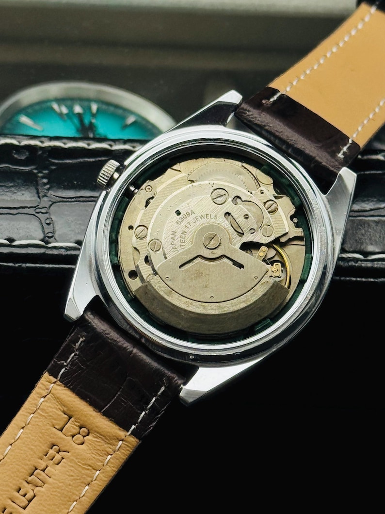 May include: Close-up of a vintage wristwatch with a silver case and a brown leather strap. The watch's back is transparent, revealing the intricate mechanical movement. The strap is marked "LEATHER 18".
