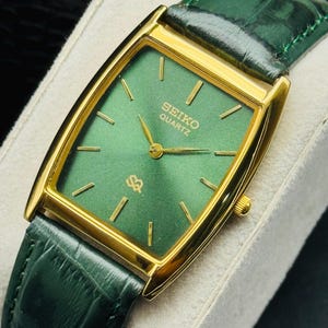 May include: A gold-toned Seiko Quartz watch with a rectangular green face and gold hands. The watch has a green leather band. The watch face displays the Seiko Quartz logo and gold hour markers.