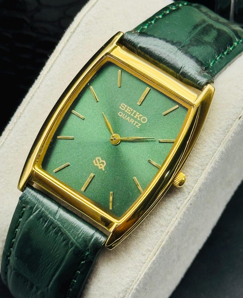 May include: A gold-toned Seiko Quartz watch with a rectangular green face and gold hands. The watch has a green leather band. The watch face displays the Seiko Quartz logo and gold hour markers.