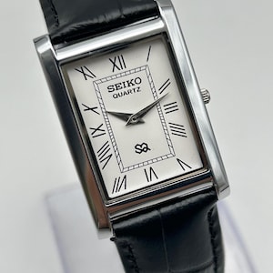 Puede incluir: Un reloj Seiko Quartz rectangular con esfera blanca y n&uacute;meros romanos negros. El reloj tiene una caja plateada y una correa de cuero negro. La esfera muestra el logotipo de Seiko Quartz y un peque&ntilde;o emblema.