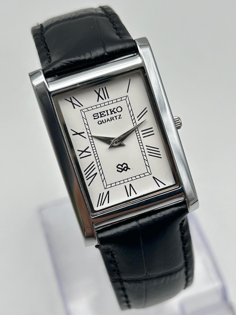 Puede incluir: Un reloj Seiko Quartz rectangular con esfera blanca y n&uacute;meros romanos negros. El reloj tiene una caja plateada y una correa de cuero negro. La esfera muestra el logotipo de Seiko Quartz y un peque&ntilde;o emblema.