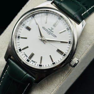 May include: A silver-toned Jaeger-LeCoultre Club watch with a white face and a dark green leather strap. The watch face features silver hands and markers, with the brand name and "CLUB" printed on the face. The strap is dark green with white stitching.