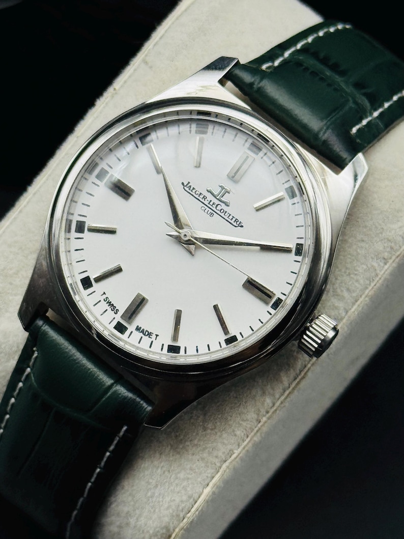 May include: A silver-toned Jaeger-LeCoultre Club watch with a white face and a dark green leather strap. The watch face features silver hands and markers, with the brand name and "CLUB" printed on the face. The strap is dark green with white stitching.