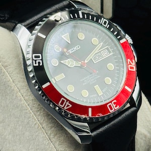 May include: A close-up of a Seiko Marine Master automatic watch with a black leather band. The watch face is gray with luminous markers and hands. The bezel is black and red, with white numerals. The watch displays "MON 15" and "JAPAN".