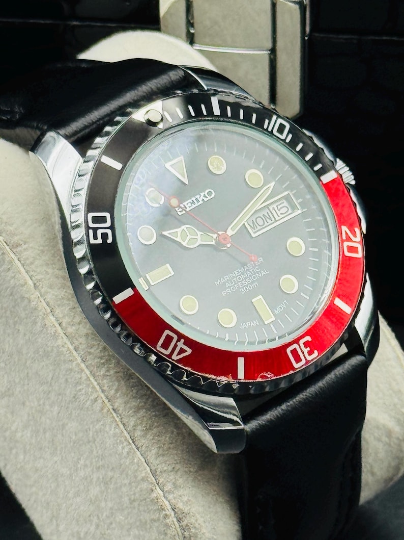 May include: A close-up of a Seiko Marine Master automatic watch with a black leather band. The watch face is gray with luminous markers and hands. The bezel is black and red, with white numerals. The watch displays "MON 15" and "JAPAN".