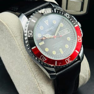 May include: A Seiko Marine Master automatic professional watch with a black leather band. The watch has a black and red bezel, a gray face, and a date display. The watch is displayed on a gray cushion.
