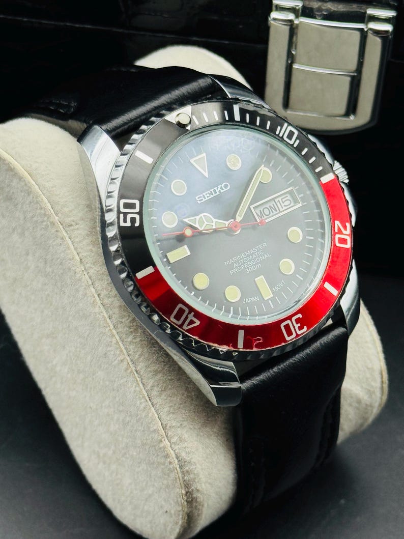 May include: A Seiko Marine Master automatic professional watch with a black leather band. The watch has a black and red bezel, a gray face, and a date display. The watch is displayed on a gray cushion.