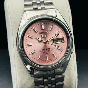 May include: A stainless steel Seiko 5 Sports watch with a pink dial. The watch features a day and date display, luminous hands, and a stainless steel bracelet. The dial reads "SEIKO 5 SPORTS WATER 10 BAR RESIST AUTOMATIC JAPAN 7009-876L R".