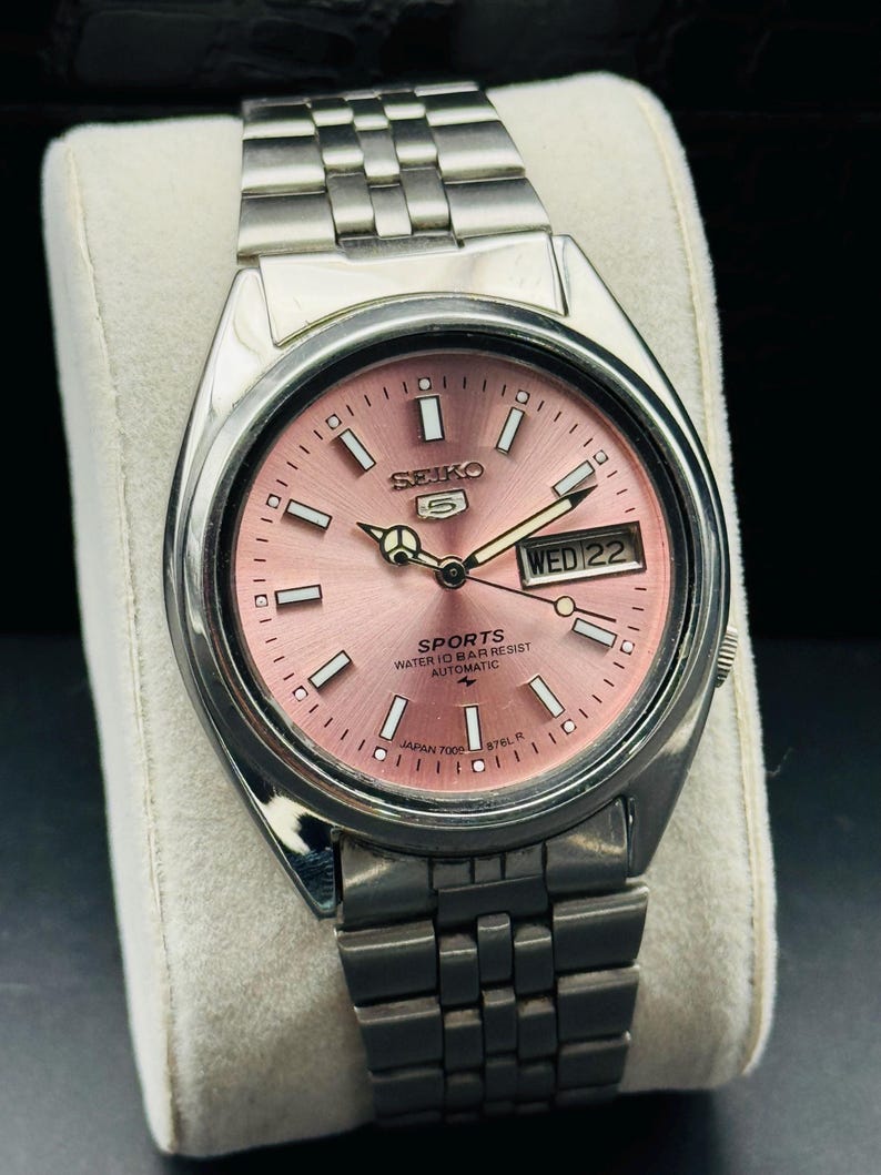 May include: A stainless steel Seiko 5 Sports watch with a pink dial. The watch features a day and date display, luminous hands, and a stainless steel bracelet. The dial reads "SEIKO 5 SPORTS WATER 10 BAR RESIST AUTOMATIC JAPAN 7009-876L R".