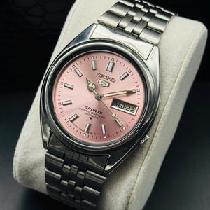 May include: A stainless steel Seiko 5 Sports automatic watch with a pink dial. The watch features a day and date display, luminous hands, and a silver-tone bracelet. The watch is displayed on a white cushion.