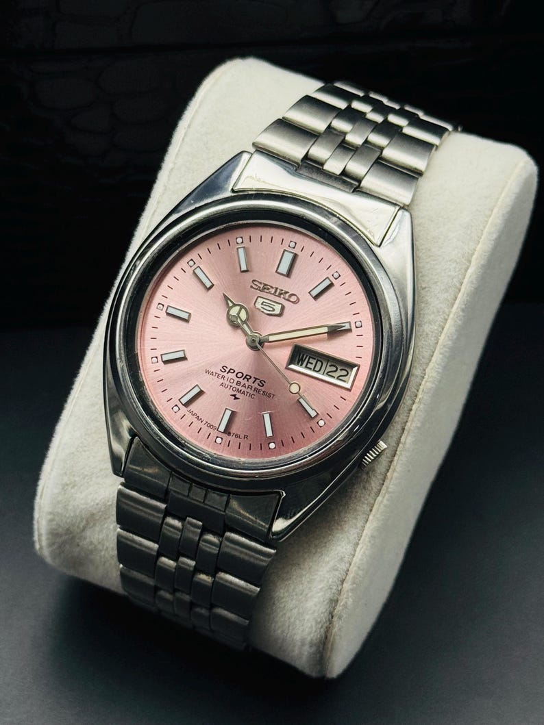 May include: A stainless steel Seiko 5 Sports automatic watch with a pink dial. The watch features a day and date display, luminous hands, and a silver-tone bracelet. The watch is displayed on a white cushion.